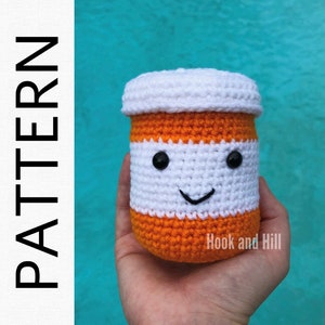 May include: A crocheted pill bottle with a white lid, orange body, and a smiling face. The bottle is held in a hand. The text "Hook and Hill" is visible on the bottle.