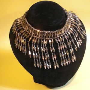 Puede incluir: Un collar de cuentas plateadas y doradas con una franja de cuentas colgantes. El collar se muestra en un busto de maniquí de terciopelo negro.