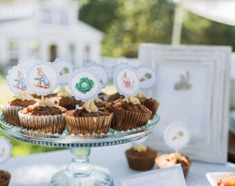 May include: A white cake stand with a glass top holds a variety of cupcakes decorated with Peter Rabbit themed toppers. The cupcakes are topped with brown frosting and have a light brown base. The stand is on a white table with a white frame picture behind it.