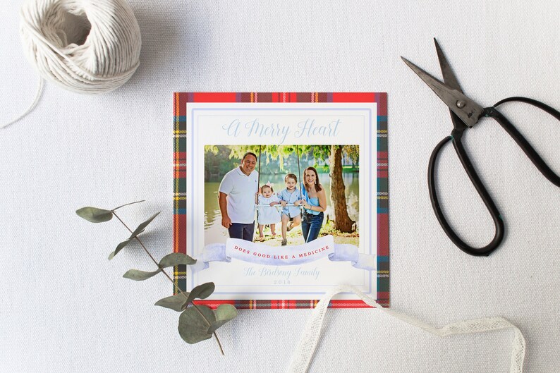 May include: A square family photo card with a plaid border in red, green, and yellow. The photo features a family of four. Text includes "A Merry Heart" and "Does good like a medicine". A ball of twine and scissors are on the white surface.