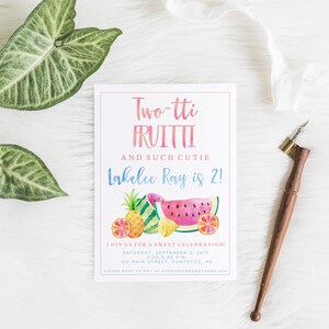 May include: A white invitation card with the text "Two-tti FRUITTI" and "Lakelee Ray is 2!" in pink and blue. The card features watercolor illustrations of fruits. A wooden calligraphy pen and green leaves are also present.