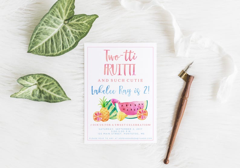 May include: A white invitation card with the text "Two-tti FRUITTI" and "Lakelee Ray is 2!" in pink and blue. The card features watercolor illustrations of fruits. A wooden calligraphy pen and green leaves are also present.