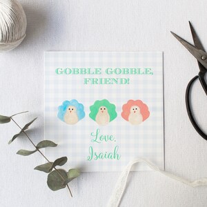 May include: A white card with a blue and white checkered pattern. The card has the text "Gobble Gobble, Friend!" and three cartoon turkeys with blue, green, and pink feathers. The card is signed "Love, Isaiah".
