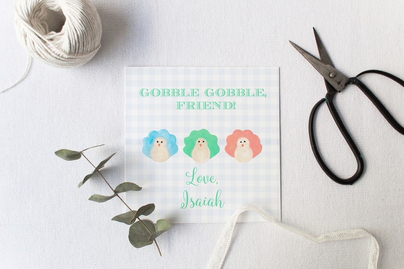 May include: A white card with a blue and white checkered pattern. The card has the text "Gobble Gobble, Friend!" and three cartoon turkeys with blue, green, and pink feathers. The card is signed "Love, Isaiah".