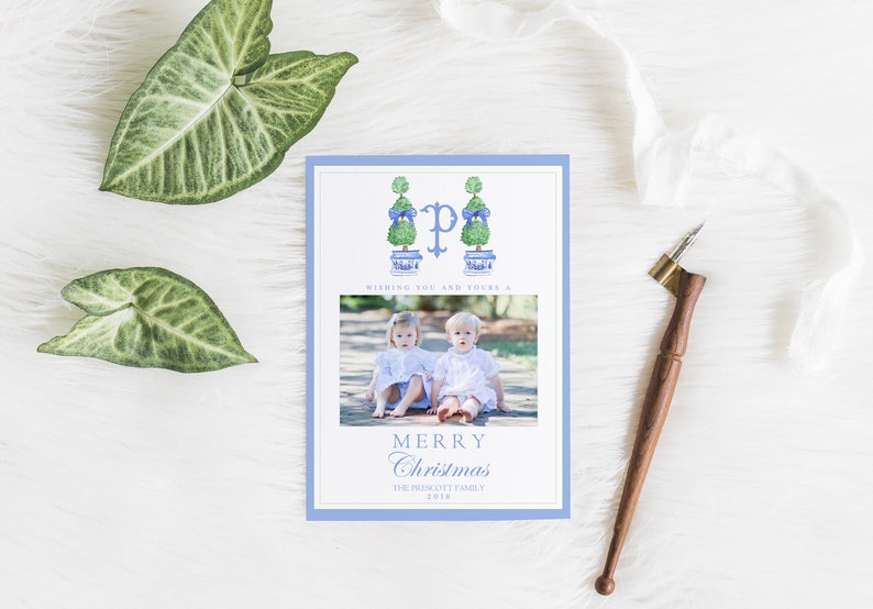Puede incluir: Una tarjeta de Navidad con un borde azul que presenta una foto de dos ni&ntilde;os peque&ntilde;os sentados en un camino. La tarjeta tiene una ilustraci&oacute;n azul y blanca de dos &aacute;rboles topiarios con lazos azules y la letra "P" en el centro. El texto "Wishing you and yours a Merry Christmas The Prescott Family 2018" est&aacute; impreso en la tarjeta.