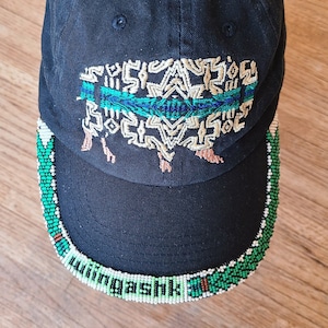 May include: Black baseball cap with a green, blue and gold embroidered design. The brim of the cap is decorated with a beaded trim in green, white and brown.