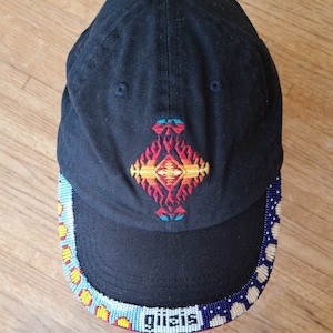 May include: Black baseball cap with a colourful geometric design embroidered on the front. The brim is trimmed with a beaded band in blue, white, yellow, and red.