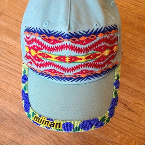 May include: A light blue baseball cap with a colorful embroidered pattern around the crown and a beaded trim around the brim. The trim features the word "miiwah" in yellow letters.
