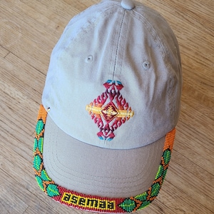 May include: A beige baseball cap with a colourful geometric design embroidered on the front. The brim is decorated with a beaded trim in shades of green, orange, red, and yellow. The trim reads "Fiesta" in yellow.