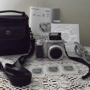 May include: A silver digital camera with a black carrying case, a USB cable, a CD, and three memory cards. The camera has a lens cover and a flash.