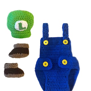 May include: A handmade crochet baby outfit in a video game theme. The set includes a green hat with a white "L", a blue romper with yellow buttons, and two pairs of brown and tan booties. The outfit is designed for a baby.