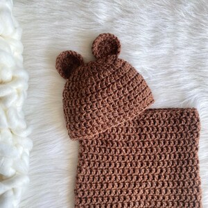 May include: A brown crocheted baby hat with bear ears and a matching brown crocheted swaddle blanket.