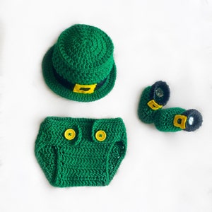 May include: Green crocheted baby outfit with a hat, diaper cover, and booties. The hat has a black band with a gold buckle. The booties have black trim and yellow buckles.