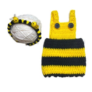 May include: A yellow and black striped crocheted baby outfit with a matching headband featuring bee accents. The outfit has a crocheted bodice with two black buttons and the headband has three felt bees.