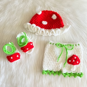 May include: A crocheted mushroom-themed baby outfit set in white and red with green accents. The set includes a hat, shorts, and booties.
