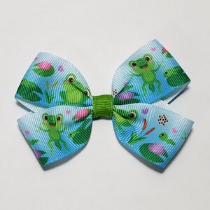 May include: A blue and white hair bow with a frog and lily pad pattern. The bow is tied with a green ribbon.