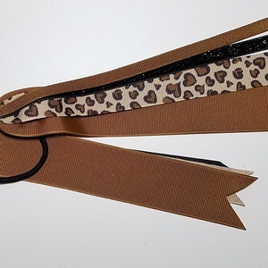 May include: A brown and black hair tie with multiple ribbons attached. The ribbons are brown, black, and a leopard print with black hearts.