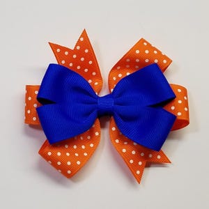 May include: A layered hair bow featuring a vibrant blue center bow atop orange ribbon with white polka dots. The bow is a cheerful accessory, perfect for adding a pop of color to any hairstyle. The bow is approximately 5 inches wide.