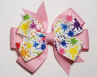 May include: A pink hair bow with a white ribbon featuring a colorful paint splatter design.