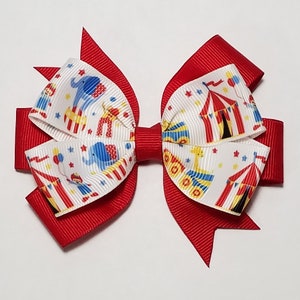 May include: A red and white hair bow with a circus theme. The bow features a white background with red and yellow circus tents, blue elephants, clowns, and stars. The bow is tied with a red ribbon.