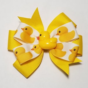 May include: Yellow hair bow with a yellow rubber ducky in the center. The bow is made of yellow ribbon with a white background and a pattern of yellow rubber ducks.