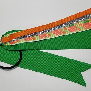 May include: A green, orange, and silver hair bow with a black elastic band. The bow features layers of green, orange, and patterned ribbons with a glitter accent. The patterned ribbon has a repeating apple design.