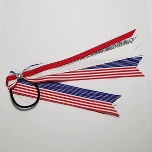 May include: A red, white, and blue hair tie with a sparkly silver ribbon and a small rhinestone bow. The hair tie is made of fabric and has a patriotic theme.