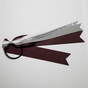 May include: A maroon, gray, white, and silver ribbon hair accessory. The hair tie features a bow with a silver accent and multiple layered ribbons. The ribbons are in shades of maroon, gray, white, and a silver glitter ribbon. The hair tie is on a white background.