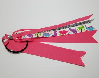 Può includere: Un elastico per capelli rosa con un nastro bianco con stampa di dinosauri, un nastro argentato glitterato e un nastro rosa.