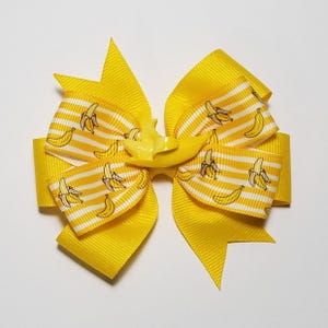 May include: A bright yellow hair bow with a banana theme. The bow features a solid yellow base with layered ribbons. Some ribbons have a white stripe pattern with banana illustrations. A yellow banana charm is in the center.