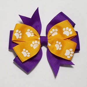 May include: A yellow bow with white paw prints on it, layered over a purple bow. The bow is made of ribbon and is suitable for hair.