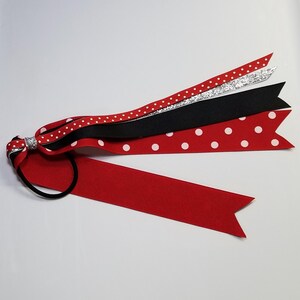 May include: A red hair tie with multiple ribbons. The ribbons include a solid red ribbon, a red ribbon with white polka dots, a black ribbon, and a silver glitter ribbon. The ribbons are attached to a black elastic band.
