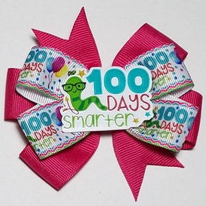 May include: A pink hair bow with a white ribbon featuring a green worm with glasses and the text "100 Days Smarter".