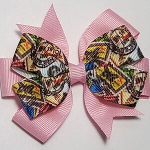 May include: A pink hair bow with a colorful patterned ribbon featuring a variety of stamps. The bow is made of grosgrain ribbon and has a simple, classic design.