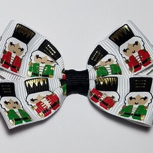 May include: A white bow with a black center, featuring a repeating pattern of nutcracker characters in red, green, gold, and black.