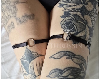 May include: A pair of black garter belts with silver metal rings. The garter belts are worn on the upper thighs of a person with tattoos.