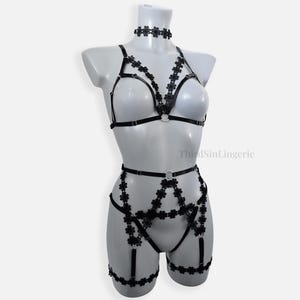 Może przedstawiać: Czarny komplet bielizny z paskami typu harness, ozdobiony małymi kwiatowymi motywami z koronki. Zestaw zawiera choker, braletkę oraz pas do pończoch z paskami na uda, połączone srebrnymi metalowymi kółkami i regulowanymi suwakami.