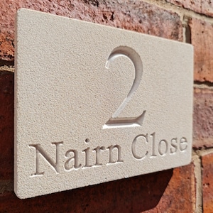 Op de afbeelding: Een rechthoekige, lichtbeige stenen huisnummerplaat met het nummer "2" en de woorden "Nairn Close" in het oppervlak gegraveerd. De plaat is bevestigd aan een rode bakstenen muur.