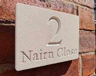 May include: A rectangular light beige stone house sign with a textured surface. The number 2 is engraved in the center, and the text Nairn Close is engraved below it in a serif font. The sign is mounted on a red brick wall.