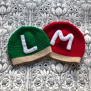 Könnte beinhalten: Zwei gehäkelte Mützen, eine grüne mit einem weißen "L" und eine rote mit einem weißen "M".