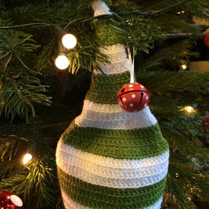 Pode incluir: Um chapéu de crochê listrado verde e branco com um pompom no topo. O chapéu está pendurado em uma árvore de Natal com enfeites vermelhos e brancos.