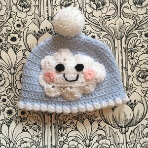 May include: A light blue crocheted hat with a white pom pom on top. The hat features a white crocheted cloud with a smiling face and pink cheeks.