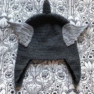 Könnte beinhalten: Graue Häkelmütze mit einer spitzen Spitze und zwei grauen Häkel Flügeln an den Seiten.