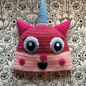 Könnte beinhalten: Rosa und blaue gehäkelte Einhornmütze mit einem weißen Horn und schwarzen Augen.