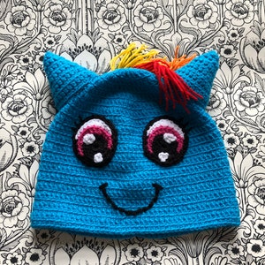 Könnte beinhalten: Eine blaue Häkelmütze mit einem Einhorn-Gesicht, einer Regenbogenmähne und schwarzen Augen. Die Mütze hat einen weißen Hintergrund für die Augen und einen rosa Umriss.