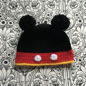 Könnte beinhalten: Schwarze Häkelmütze mit rotem und gelbem Besatz und weißen Bommeln. Die Mütze hat zwei weiße Kreise auf der Vorderseite, die an Mickey Mouse-Ohren erinnern.