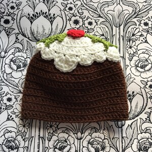 May include: A brown crocheted hat shaped like a Christmas pudding with green and white crocheted details and a red pom-pom on top.