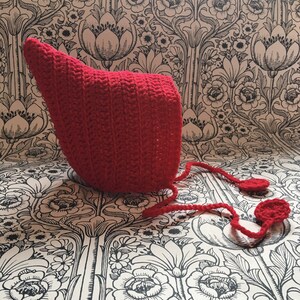 May include: A red crocheted baby bonnet with a long tie. The bonnet is made with a simple stitch and has a pointed shape.