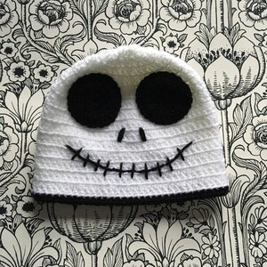 Könnte beinhalten: Weiße Häkelmütze mit schwarzen Details, inspiriert von Jack Skellington. Die Mütze hat schwarze Augen, ein gesticktes Lächeln und einen schwarzen Rand.