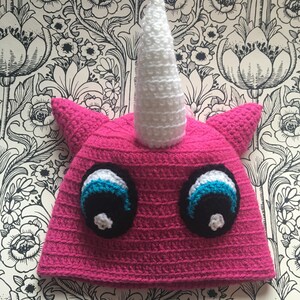 Könnte beinhalten: Rosa gestrickte Einhornmütze mit weißem Horn und schwarzen und blauen Augen.
